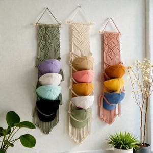 Op de afbeelding: Drie macramé hoedenorganizers in groen, crème en roze, elk met een verscheidenheid aan baseballpetten in verschillende kleuren. De organizers hangen aan een witte muur, met een plant op de voorgrond.
