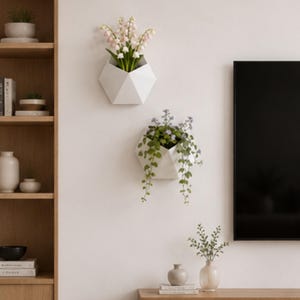 May include: Two white geometric wall planters, each holding greenery and flowers. One planter contains pink flowers, the other has trailing green plants with small purple flowers. The planters are mounted on a white wall.