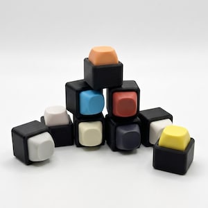 Può includere: Una collezione di oggetti quadrati colorati. Ogni oggetto ha una parte superiore colorata e una base nera. I colori includono arancione, blu, rosso, bianco, giallo e grigio. Gli oggetti sono disposti a forma di piramide.