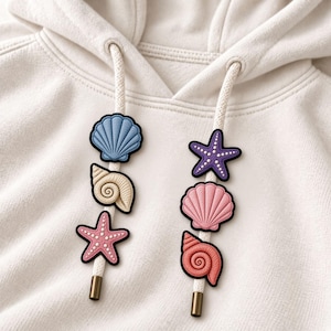 Puede incluir: Una sudadera con capucha color crema con dos cordones blancos adornados con parches bordados de vida marina. El cordón izquierdo tiene una concha azul, una concha espiral beige y una estrella de mar rosa. El cordón derecho tiene una estrella de mar morada, una concha rosa y una concha espiral coral.