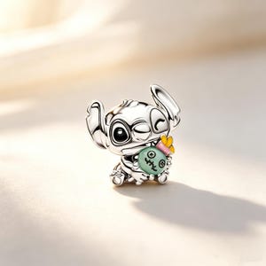 Puede incluir: Un charm de plata que representa al personaje Stitch de la pel&iacute;cula animada Lilo & Stitch. El charm gui&ntilde;a un ojo y sostiene una bola verde con una cara, acentuada con un lazo rosa y una flor amarilla.