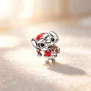 Puede incluir: Un charm de plata con un personaje de dibujos animados que lleva un gorro y una chaqueta rojos de Pap&aacute; Noel, sosteniendo un hombre de jengibre. El charm tiene ojos negros y est&aacute; sobre un fondo claro borroso.