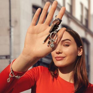 May include: A silver snake chain bracelet with a heart-shaped clasp and a dangling heart charm, paired with a silver key ring featuring a circular charm with a heart cutout and a small dangling heart, held up against a blurred urban background.