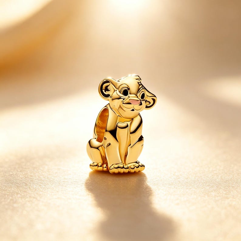 Puede incluir: Un charm dorado que representa un cachorro de le&oacute;n sentado, con una nariz rosada y orejas peque&ntilde;as y redondeadas. El charm es muy detallado y tiene un acabado pulido, lo que sugiere que est&aacute; hecho de metal. El cachorro est&aacute; sentado erguido, con sus patas visibles.