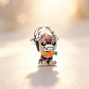 Puede incluir: Un charm plateado con forma de reno, sosteniendo una zanahoria naranja con la parte superior verde. El reno tiene detalles marrones y blancos, con cuernos plateados y un cuerpo moteado marr&oacute;n rojizo. El charm es para un brazalete o collar.