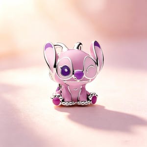 Puede incluir: Un charm rosa y morado que representa un personaje de dibujos animados con orejas grandes y una expresi&oacute;n gui&ntilde;ando un ojo. El charm tiene un contorno plateado y est&aacute; sobre un fondo rosa suave. El personaje tiene ojos grandes, una nariz peque&ntilde;a y una boca sonriente.