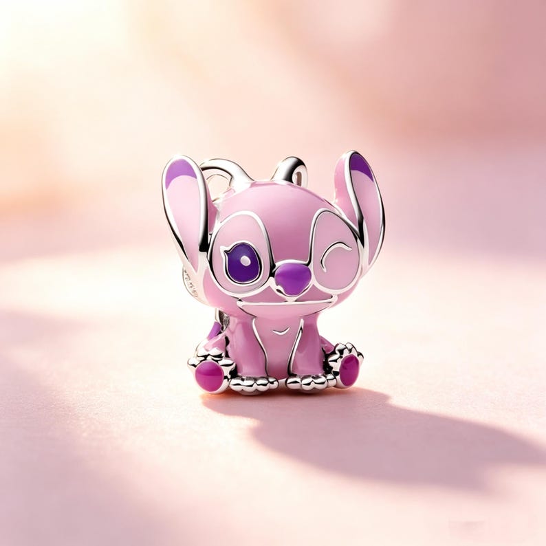 Puede incluir: Un charm rosa y morado que representa un personaje de dibujos animados con orejas grandes y una expresi&oacute;n gui&ntilde;ando un ojo. El charm tiene un contorno plateado y est&aacute; sobre un fondo rosa suave. El personaje tiene ojos grandes, una nariz peque&ntilde;a y una boca sonriente.