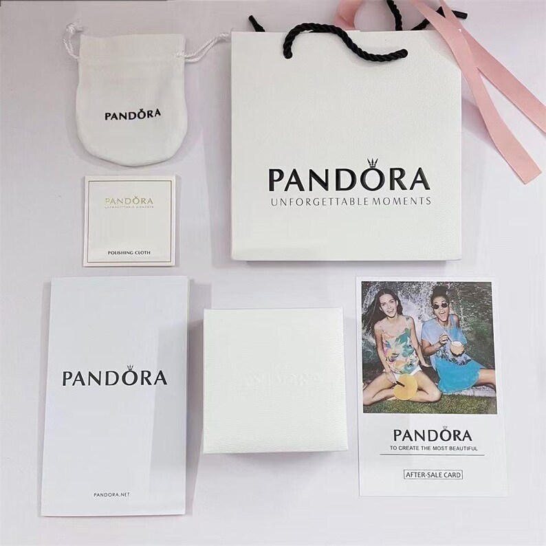 Puede incluir: Una colecci&oacute;n de art&iacute;culos de la marca Pandora, que incluye una bolsa de regalo blanca con asas negras, una peque&ntilde;a bolsa, un pa&ntilde;o de pulido, una caja y una tarjeta con dos mujeres. El logotipo de Pandora es visible.