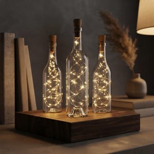 Puede incluir: Tres botellas de vidrio transparente con tapones de corcho, cada una con una cadena de luces LED de color blanco cálido. Las botellas están dispuestas sobre una base rectangular de madera oscura, creando un efecto luminoso con cables visibles.