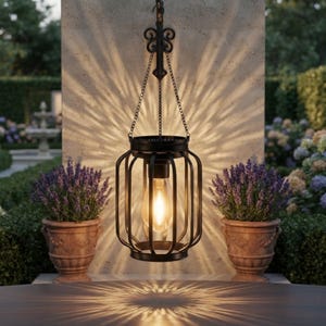 May include: A black metal hanging lantern with a cage-like frame and a central glowing light bulb. The lantern is suspended by a decorative bracket and chains, casting intricate, starburst-like light patterns onto the wall behind it.