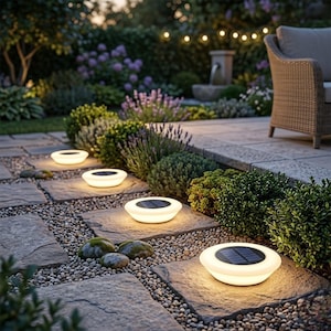 May include: Solar-powered pathway lights illuminate a stone walkway in a garden. The lights are white, circular, and have a solar panel on top. The garden features lush greenery, flowers, and a seating area, creating a serene outdoor ambiance.