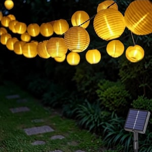 May include: A decorative string of glowing yellow paper lanterns, ideal for outdoor use. The round lanterns are suspended above a stone path and lush greenery. A solar panel is visible, indicating an eco-friendly lighting system for gardens or patios.