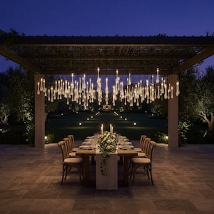 Peut inclure: Une longue table à manger en bois avec des chaises disposées sous une pergola sombre. Au-dessus de la table est suspendue une grande installation de nombreuses lampes suspendues en verre transparent en forme de goutte d'eau.