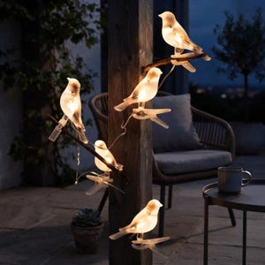 May include: A string of five illuminated bird-shaped lights, each featuring a translucent white body that glows with a warm light. The birds are attached to clips and are positioned along a dark wooden post, creating a decorative lighting display.