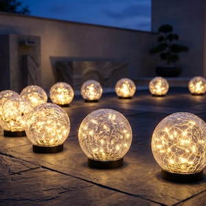 Puede incluir: Una colección de luces de jardín esféricas de vidrio con textura agrietada y luces de cadena de color blanco cálido en su interior. Cada globo se asienta sobre una base negra circular, emitiendo un brillo suave sobre la piedra oscura.