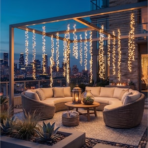 May include: A curved wicker outdoor sofa set with beige cushions arranged around a round wooden coffee table. A metal lantern and a glass bowl sit on the table. Strings of warm white fairy lights hang from the overhead pergola structure.