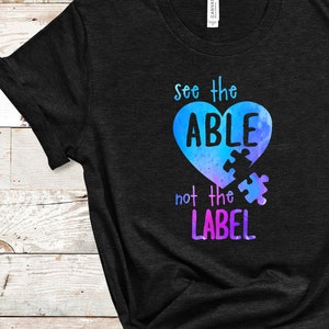 Może przedstawiać: Czarny T-shirt z niebiesko-fioletowym sercem, na którym widnieje napis "see the ABLE not the LABEL" z puzzlem w sercu.