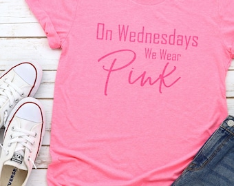 May include: Pink t-shirt with the text "On Wednesdays We Wear Pink" in a white font. The shirt is laying on a white wooden surface with a pair of white sneakers and a pair of blue denim shorts.