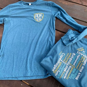 Op de afbeelding: Twee t-shirts met lange mouwen in een tealblauwe kleur. De voorkant van het ene shirt heeft een geel en zwart logo met de tekst "ADK TUG HILL". De achterkant van het andere shirt heeft een woordwolk met de namen van steden in de Adirondack-regio van New York.