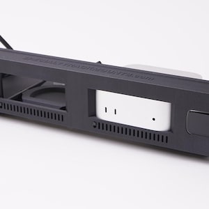 May include: A black rack mount with multiple openings and a white component. The rack mount has a rectangular shape with ventilation slots and a small, square button. The text "ULTIMATE MACMOUNTS.COM" is printed on the top.
