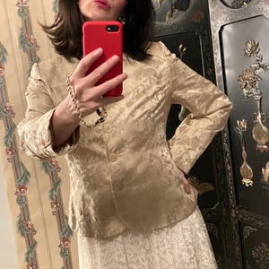 May include: A gold-colored blazer with a floral pattern. The blazer has three buttons and long sleeves. The person in the image is wearing a white lace skirt. The person is holding a red phone.