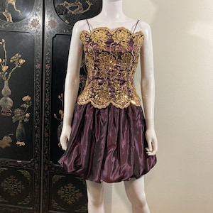 May include: A short, purple dress with a gold embroidered bodice. The bodice has a scalloped neckline and is adorned with sequins. The skirt is puffy and has a gathered look.