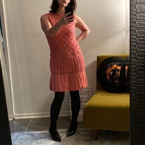 May include: A pink crocheted dress with a scalloped hemline. The dress is sleeveless and has a fitted bodice. The model is wearing black tights and black heels.