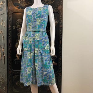 May include: A sleeveless vintage dress in a blue, green, and white paisley pattern. The dress features a square neckline, a fitted bodice, a matching belt, and two front pockets. The dress is displayed on a mannequin.