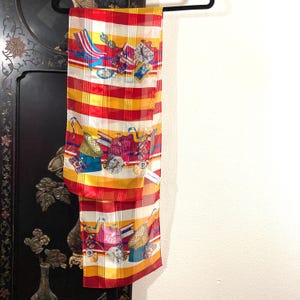 May include: A red, yellow, and white striped silk scarf with a colorful print of shopping bags and hats. The scarf has a white tag with the text "Made in Italy".