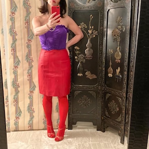 May include: A strapless purple corset top with a ruffled neckline paired with a red pencil skirt and matching red tights. The outfit is completed with red strappy heels. A black decorative screen is in the background.