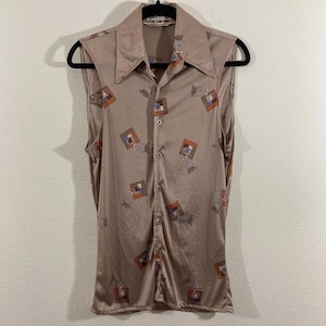 May include: A sleeveless button-down shirt in a tan color, featuring a large collar and a pattern of geometric shapes in shades of brown, orange, and blue. The shirt is hanging on a black hanger against a plain white background.