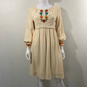 May include: Cream-colored vintage dress with long sleeves and a knee-length hem. The dress features colorful floral embroidery around the neckline and cuffs, and a braided belt detail at the waist. The dress is displayed on a mannequin.