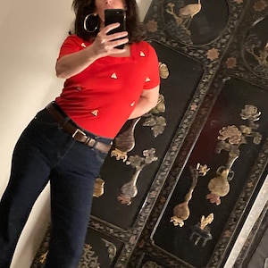 May include: A woman wearing a red short-sleeved shirt with a geometric pattern and dark blue jeans. She is standing in front of a black lacquered screen with gold accents.
