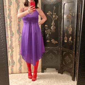 May include: A purple spaghetti strap dress with a flowing, asymmetrical hemline. The dress is paired with red tights and red strappy heels. The background includes a decorative folding screen and a floral patterned wall.