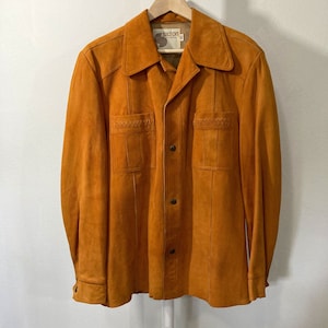 May include: A brown suede jacket with a button-up front and long sleeves. The jacket has two flap pockets on the front and a woven detail on the pockets. The label on the jacket reads "Silton".