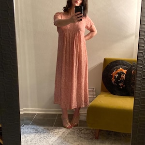 May include: A pink maxi dress with a floral print. The dress has a fitted bodice and a flowing skirt. The model is wearing the dress with sandals.