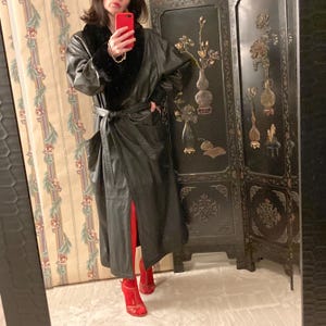 May include: Full-length black leather coat with a black fur collar and a matching belt. The coat is worn with red heels and red tights. The background includes a floral patterned wallpaper and a black decorative screen.