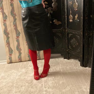 May include: A black leather skirt paired with red tights and red high-heeled sandals. The outfit is completed with a turquoise top. The background includes a decorative screen and a patterned wall.