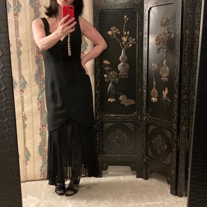 May include: A black sleeveless dress with a V-neckline and a layered skirt. The dress has a fitted bodice and a flowing, sheer lower section. The dress is paired with patterned stockings. A decorative folding screen is in the background.