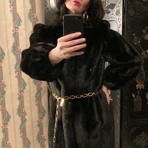 May include: A dark brown faux fur coat with long sleeves and a gold chain belt. The coat has a plush texture and a classic design. The gold belt adds a touch of elegance to the overall look.