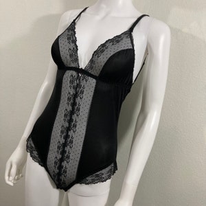May include: Black bodysuit with a plunging neckline and delicate spaghetti straps. The bodice features a sheer, dotted panel with floral lace detailing, and is trimmed with lace along the neckline and leg openings. The garment is displayed on a mannequin.