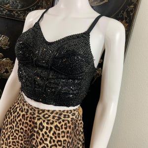 May include: A black sequined crop top with thin straps. The top is heavily embellished with sequins and beads, creating a textured surface. The top is paired with a leopard print skirt.