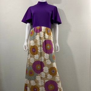 May include: A vintage maxi dress with a purple bodice and a floral print skirt. The top features a high neckline and short, flared sleeves. The skirt has a beige background with large, stylized flowers in purple, yellow, and gray.