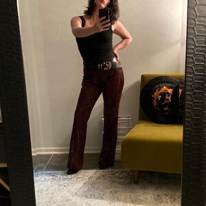May include: Burgundy velvet pants with a high waist and flared legs. The pants are paired with a black tank top and a wide black belt with a gold buckle. The outfit is reflected in a large mirror.