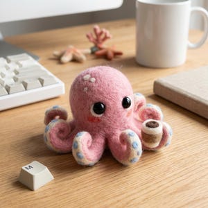 Puede incluir: Un pulpo rosa de fieltro hecho a mano con ojos negros y una pequeña taza de café. El pulpo tiene detalles azules y blancos en sus tentáculos y puntos blancos. Un accesorio de escritorio caprichoso.