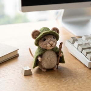 May include: A handmade felt mouse figure, dressed in a green cape and hat, with a wooden staff. The mouse has tan fur and brown ears. The figurine is on a wooden desk near a keyboard and a book.
