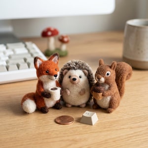 Op de afbeelding: Een grillige scène met drie vilten dierenfiguren: een vos met een kopje, een egel en een eekhoorn met een eikel. Een munt en een toetsenbordtoets liggen ervoor. Twee paddenstoelbeeldjes staan op de achtergrond.