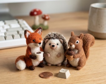 May include: A whimsical scene featuring three felted animal figurines: a fox holding a cup, a hedgehog, and a squirrel with an acorn. A penny and a keyboard key are in front of them. Two mushroom figurines are in the background.