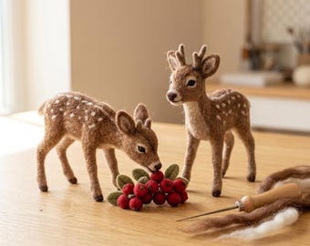 Può includere: Due statuette di cervo in lana infeltrita marrone con macchie bianche, una in piedi e una china verso un grappolo di bacche rosse in feltro con foglie verdi. Un ago da infeltrimento con manico in legno e fibre di lana sono vicini.