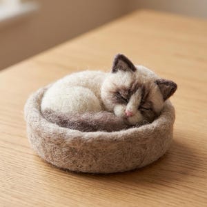 May include: A needle-felted Siamese cat figurine curled up in a round, textured beige wool bed. The cat has cream-colored fur, dark brown ears and face markings, and a small pink nose. The entire piece is crafted from soft, matted wool fibers.